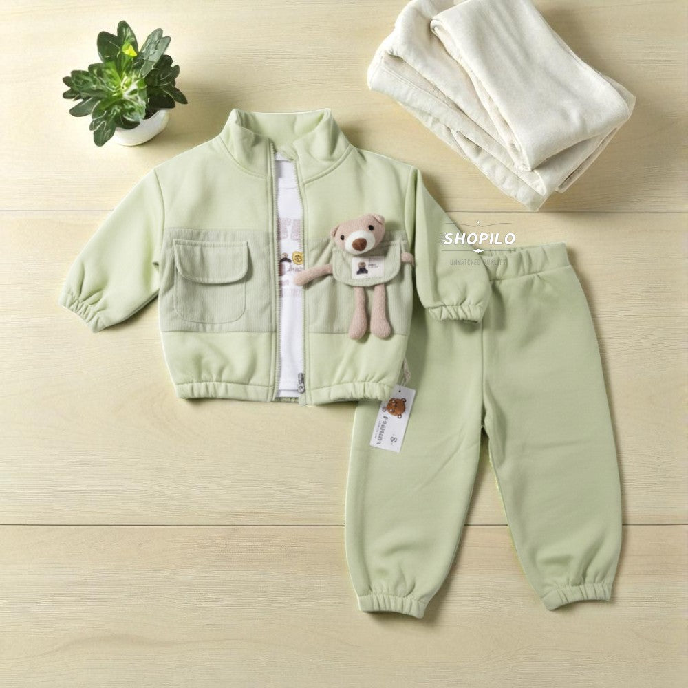 A baby two-piece clothing set in pale sage green, featuring a zip-up jacket with a teddy bear applique and matching jogger pants, laid flat on a light wood background with a small potted plant and folded cream blanket.