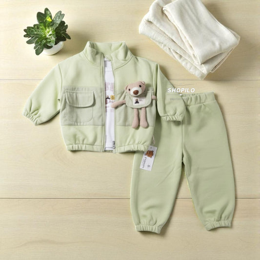 A baby two-piece clothing set in pale sage green, featuring a zip-up jacket with a teddy bear applique and matching jogger pants, laid flat on a light wood background with a small potted plant and folded cream blanket.