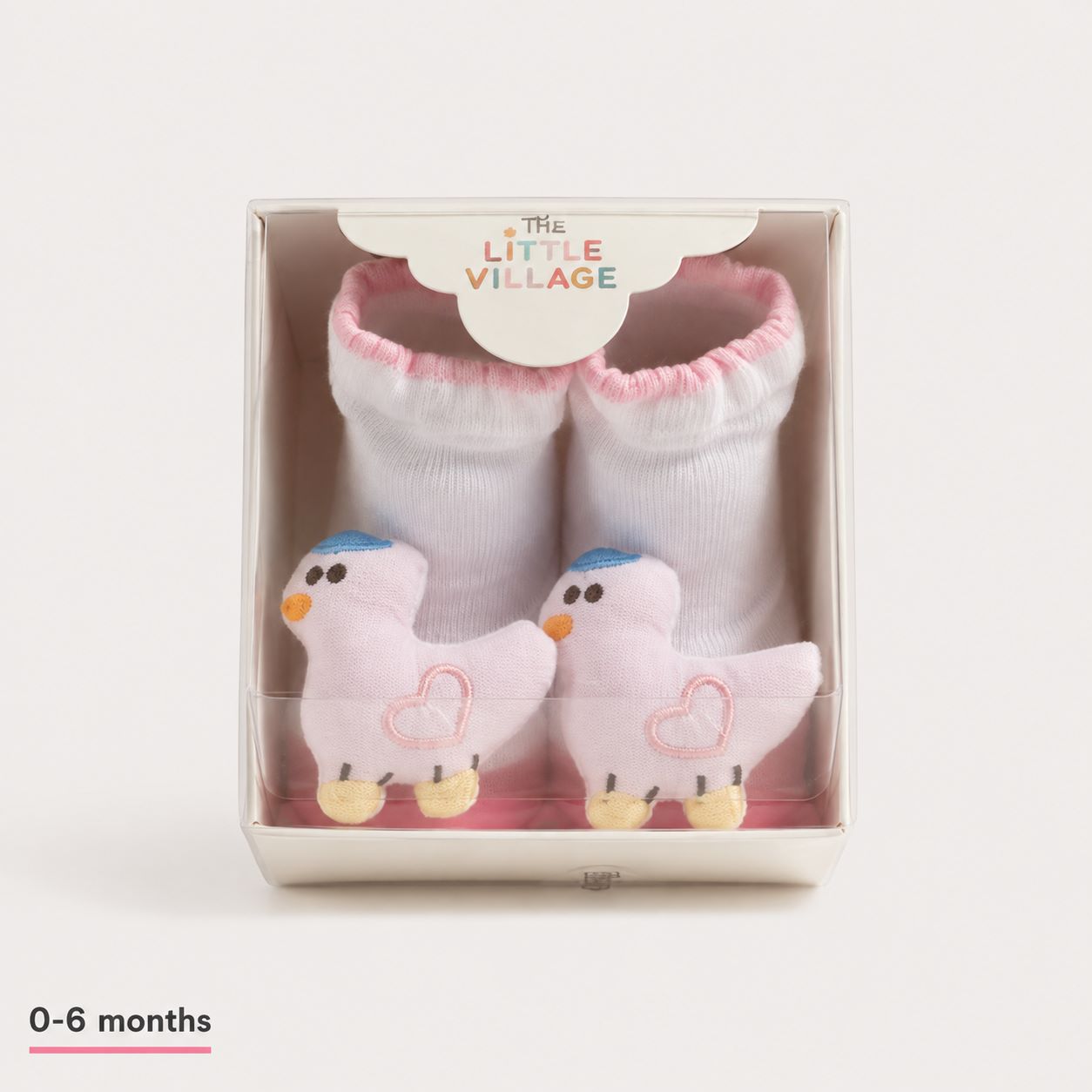A pair of baby booties by The Little Village, featuring white knit socks with pink trim and plush pink duck toys inside a clear-fronted display box. The box has a white paper insert at the top with the brand name 'THE LITTLE VILLAGE' and a '0-6 months' size label is printed in the bottom-left corner.