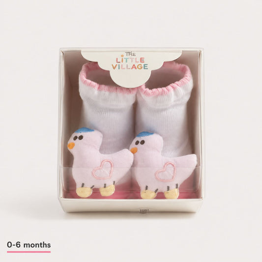 A pair of baby booties by The Little Village, featuring white knit socks with pink trim and plush pink duck toys inside a clear-fronted display box. The box has a white paper insert at the top with the brand name 'THE LITTLE VILLAGE' and a '0-6 months' size label is printed in the bottom-left corner.
