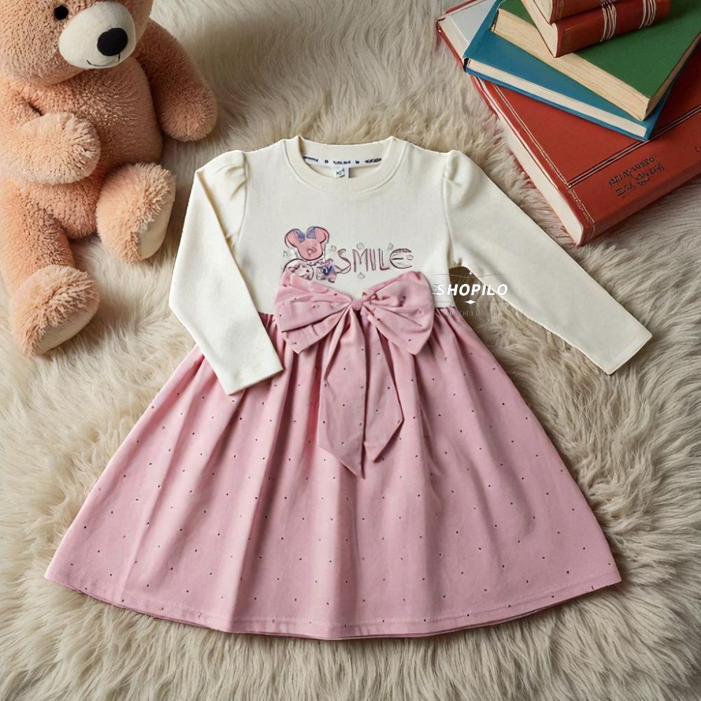 A baby girl's long-sleeve dress with a cream-colored top featuring a cartoon mouse and the word 'SMILE', a pink waistband with a large pink bow, and a pink skirt with tiny polka dots. The dress is displayed on a white faux fur rug next to a teddy bear and a stack of books.