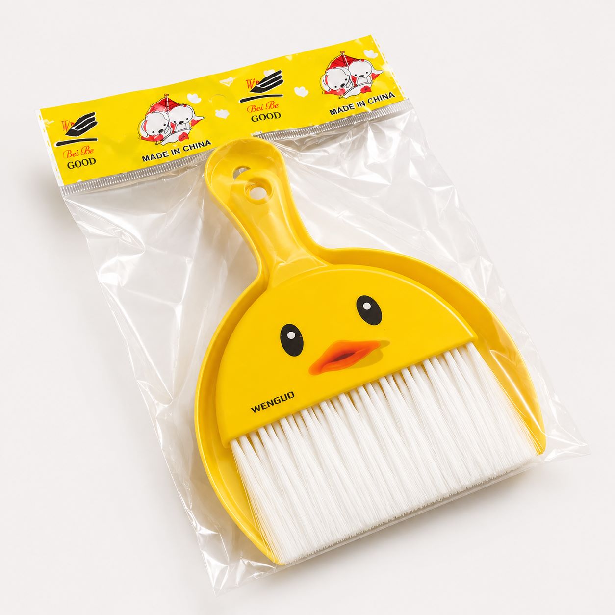 A yellow duck-shaped dustpan and brush set in clear plastic packaging with a yellow header card. The dustpan has a smiling duck face and the word 'WENGQUO' printed near the brush bristles. Cute Duck Mini Dustpan & Brush Duster Set
