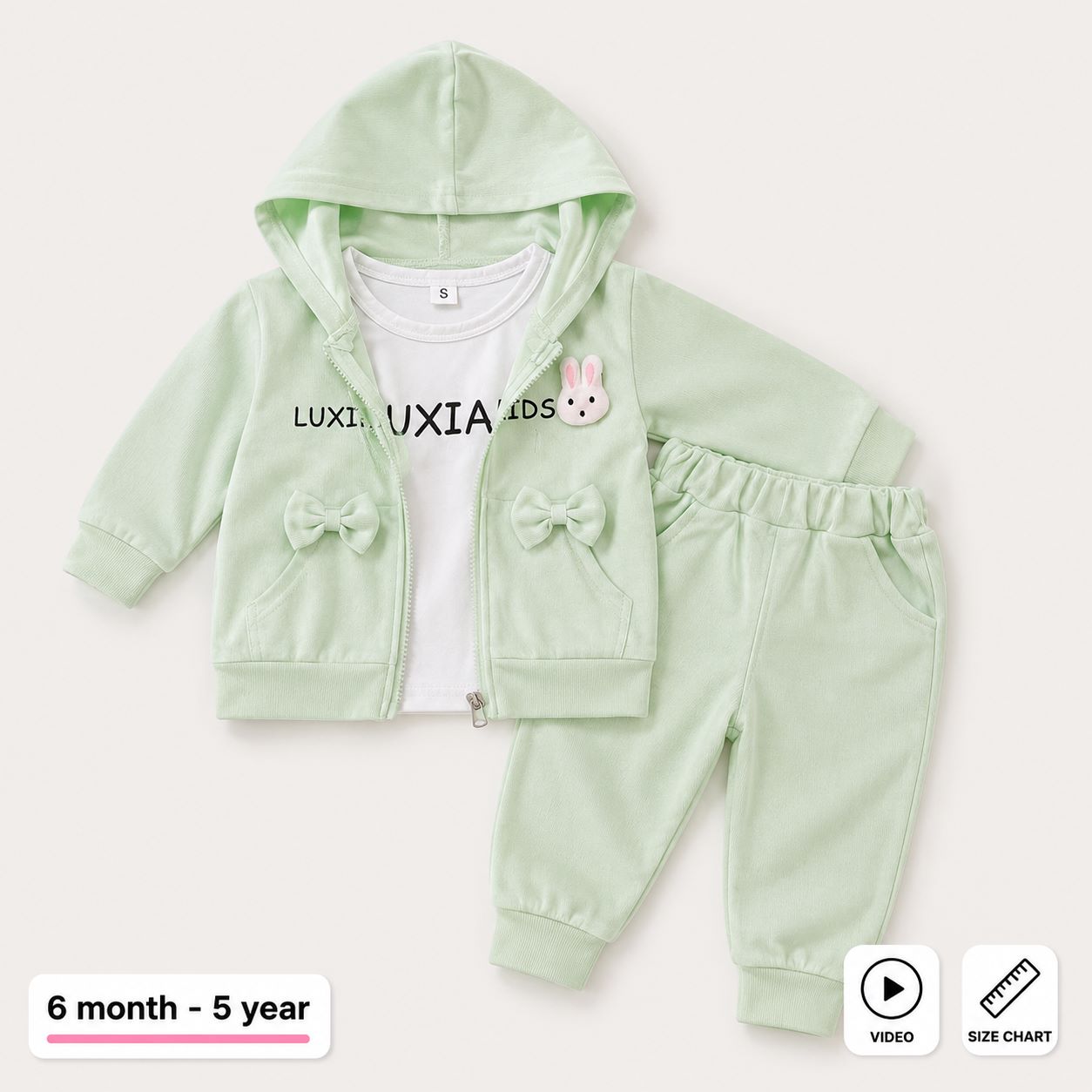 A baby girl's two-piece tracksuit set in pale mint green, featuring a zip-up hoodie with bow details and a white t-shirt underneath, shown on a light beige background with age and informational overlays.