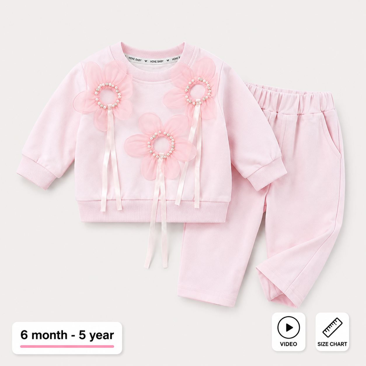 A baby girl's two-piece tracksuit in light pink, featuring a long-sleeve top with three large flower appliques and matching elastic-waist pants. The image is set against a light beige background with informational overlays indicating the age range of 6 month - 5 year, a video view, and a size chart.