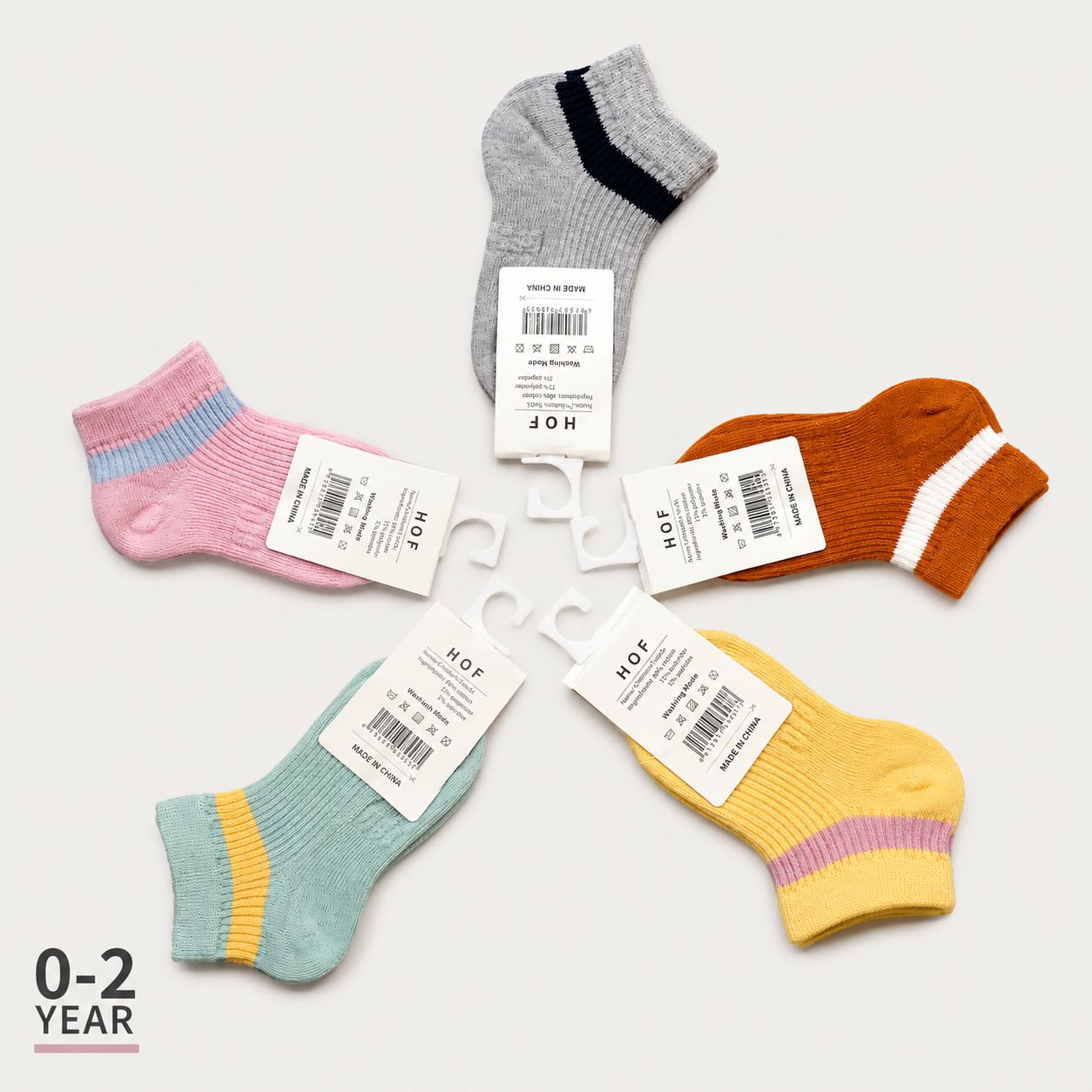 Six pairs of children's ankle socks arranged in a circle on a light gray background, each with a white paper tag. The socks come in pastel pink, mint green, yellow, orange, and heather gray, with striped accents in navy, purple, and yellow.