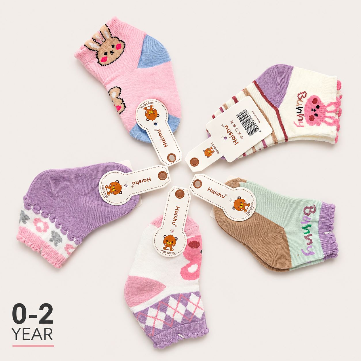 Six pairs of children's socks arranged in a circle on a light beige background, each with a bone-shaped paper tag in the center. The socks feature various pastel colors and cute cartoon animal prints, including a bunny, a pink bear, and a rabbit argyle pattern.